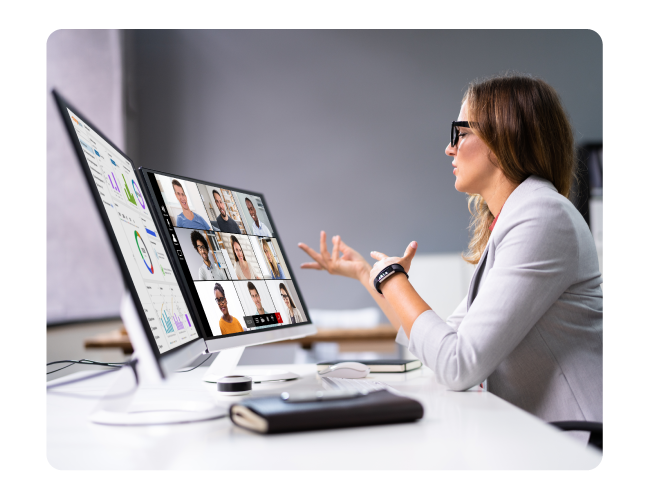 A professional at a desk speaks and gestures during a video meeting shown on a large monitor with multiple participants and data charts.
